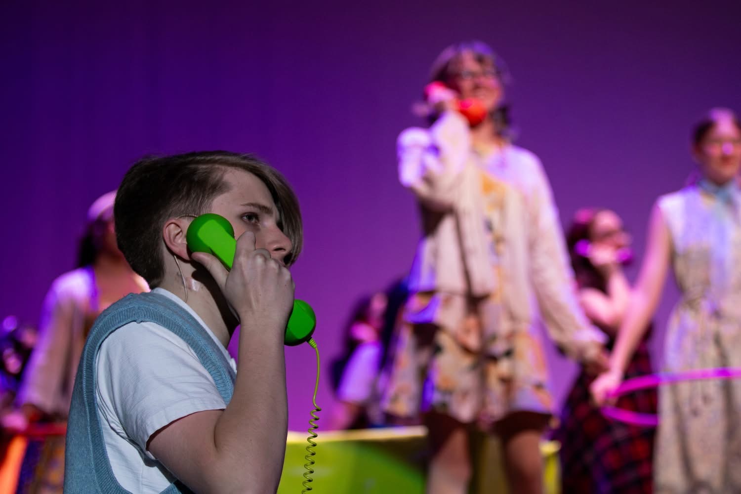 Chase performing on stage during a musical, holding a bright green phone prop under colorful stage lighting.