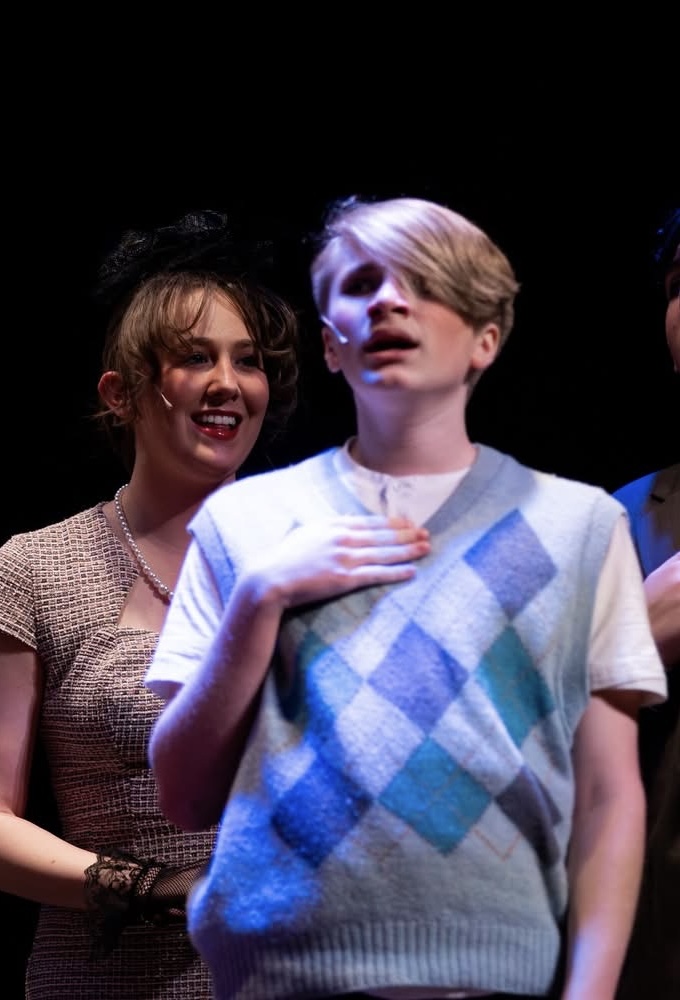 Chase singing on stage during a choir performance, hand over his chest under stage lights.